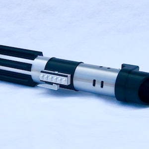 May include: A black and silver lightsaber toy with a black handle and a silver blade. The handle has a black and silver striped design and a small silver button on the side. The blade is made of plastic and has a black tip.