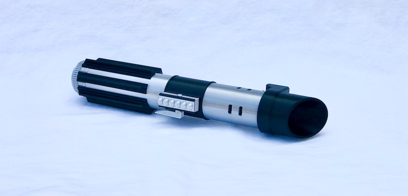 May include: A black and silver lightsaber toy with a black handle and a silver blade. The handle has a black and silver striped design and a small silver button on the side. The blade is made of plastic and has a black tip.