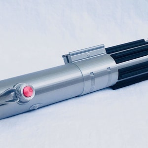 Puede incluir: Un sable de luz plateado y negro con un bot&oacute;n rojo y una empu&ntilde;adura detallada. El accesorio tiene un cuerpo cil&iacute;ndrico con una serie de aletas negras en la parte superior. El sable de luz es una r&eacute;plica de un arma de ciencia ficci&oacute;n popular.