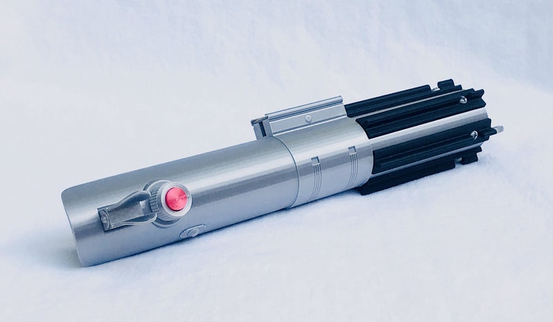 Puede incluir: Un sable de luz plateado y negro con un bot&oacute;n rojo y una empu&ntilde;adura detallada. El accesorio tiene un cuerpo cil&iacute;ndrico con una serie de aletas negras en la parte superior. El sable de luz es una r&eacute;plica de un arma de ciencia ficci&oacute;n popular.