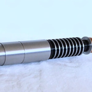 May include: A silver lightsaber prop with a black and silver blade, a copper coloured connector, and a silver handle with a knurled end.