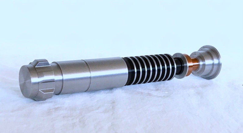 May include: A silver lightsaber prop with a black and silver blade, a copper coloured connector, and a silver handle with a knurled end.