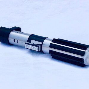 May include: A silver and black lightsaber toy with a black handle and a silver blade. The handle has a silver button and a black grip. The blade is made of plastic and has a silver finish.
