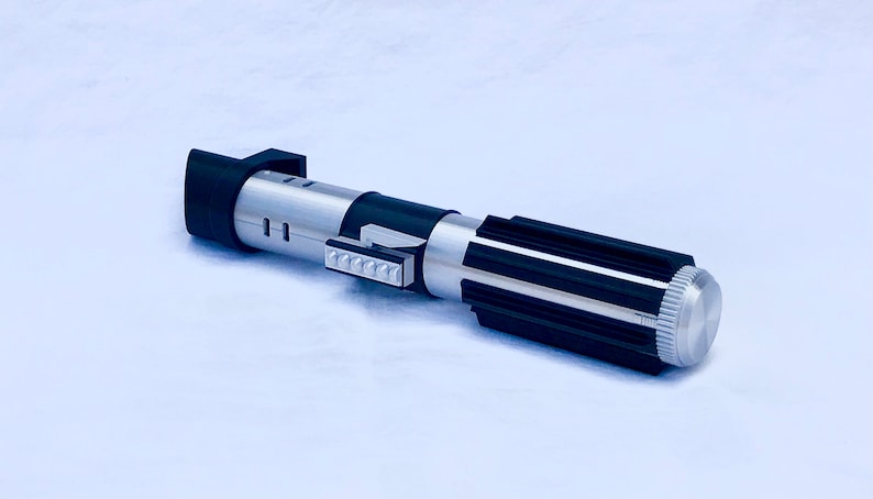 May include: A silver and black lightsaber toy with a black handle and a silver blade. The handle has a silver button and a black grip. The blade is made of plastic and has a silver finish.