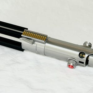 May include: A silver and black lightsaber prop with a red button and gold accents. The prop has a cylindrical body with detailed mechanical components and a black ribbed section at the top.