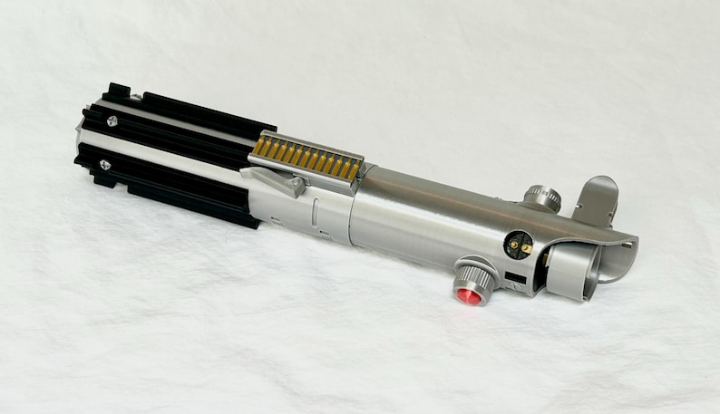 May include: A silver and black lightsaber prop with a red button and gold accents. The prop has a cylindrical body with detailed mechanical components and a black ribbed section at the top.