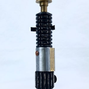May include: A black and silver lightsaber replica with a brass accent and a black base. The base has a blue label that reads "OBI-WAN KENOBI".