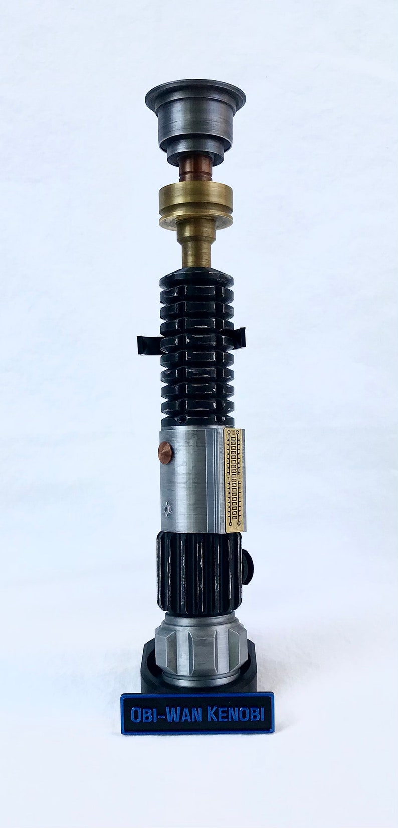 May include: A black and silver lightsaber replica with a brass accent and a black base. The base has a blue label that reads "OBI-WAN KENOBI".