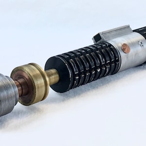 May include: A disassembled lightsaber with a silver and black handle, a brass connector, and a silver emitter. The lightsaber is on a white surface.