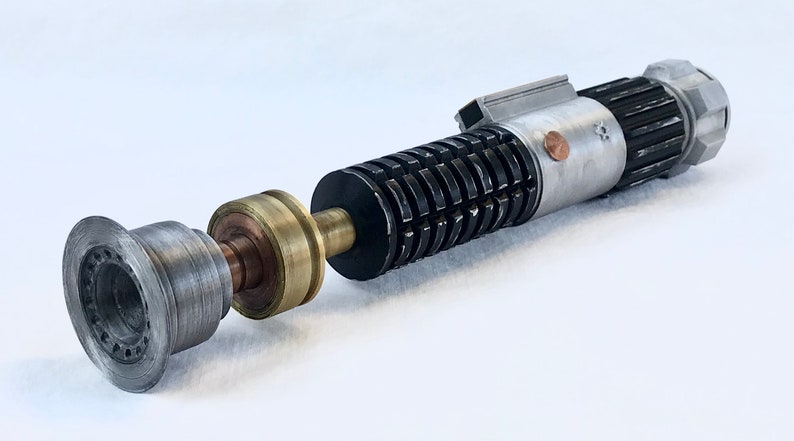 May include: A disassembled lightsaber with a silver and black handle, a brass connector, and a silver emitter. The lightsaber is on a white surface.