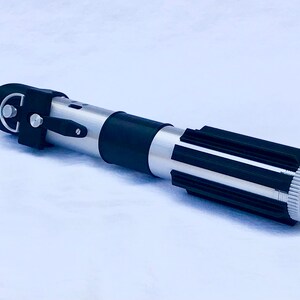 May include: A silver and black lightsaber prop with a black handle and a silver blade. The handle has a black grip and a silver knob on the end.