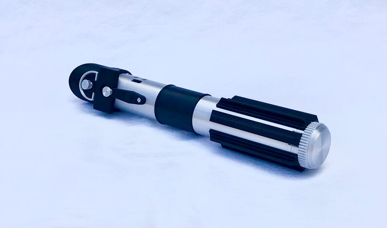 May include: A silver and black lightsaber prop with a black handle and a silver blade. The handle has a black grip and a silver knob on the end.