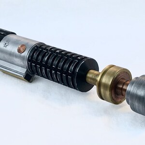 May include: A disassembled lightsaber with a silver and black body, a brass connector, and a grey emitter. The lightsaber is made of metal and has a detailed design.