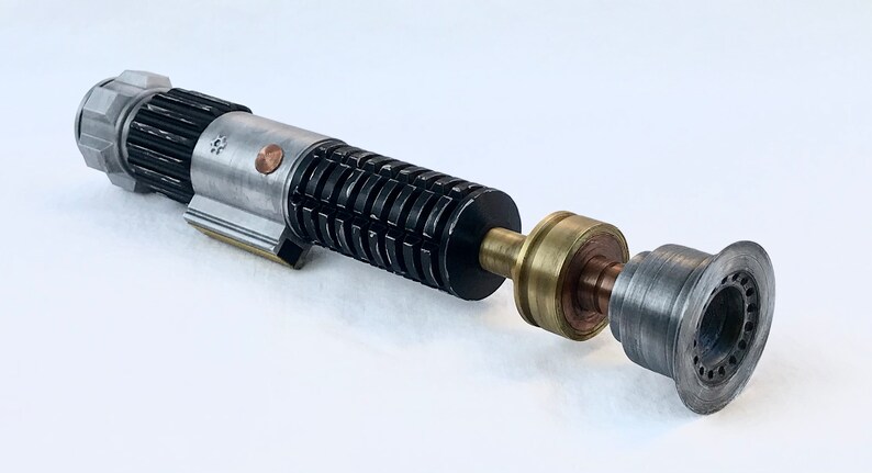 May include: A disassembled lightsaber with a silver and black body, a brass connector, and a grey emitter. The lightsaber is made of metal and has a detailed design.