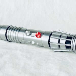 Könnte beinhalten: Ein silbernes Lichtschwert mit roten Knöpfen und einem schwarzen und silbernen Emitter. Das Lichtschwert ist aus Metall und hat eine strukturierte Oberfläche.
