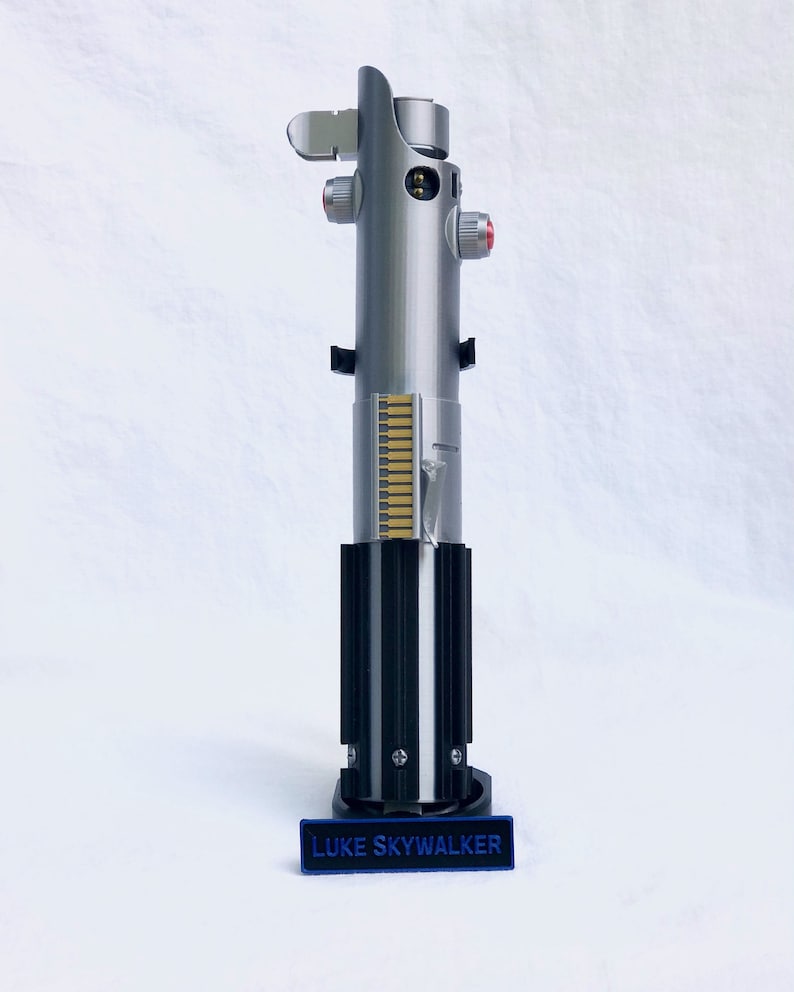 Puede incluir: R&eacute;plica de sable de luz plateado y negro con botones rojos y una base negra. El sable de luz tiene un dise&ntilde;o detallado con una serie de l&iacute;neas verticales doradas. Una placa azul en la base dice "LUKE SKYWALKER".