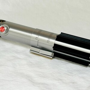 May include: A silver and black lightsaber prop with a red button and detailed design. The prop is lying on a white surface. The lightsaber has a cylindrical body with black accents.