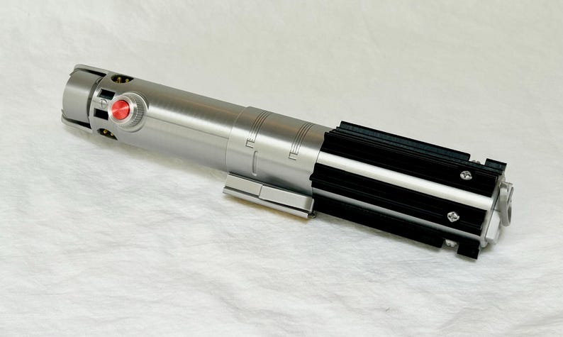 May include: A silver and black lightsaber prop with a red button and detailed design. The prop is lying on a white surface. The lightsaber has a cylindrical body with black accents.