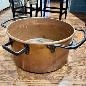 Könnte beinhalten: Ein großer Kupfertopf mit zwei schwarzen Metallgriffen. Der Topf hat eine breite, offene Oberseite und ist mit kleinen Metallnieten vernietet. Das Kupfer hat eine warme, gealterte Patina. Ein klassischer Küchenartikel.