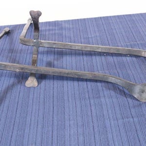 Op de afbeelding: Twee metalen trivets met een rustieke, handgemaakte uitstraling. Elke trivet heeft een gebogen, staafachtig ontwerp met hartvormige accenten aan de uiteinden en aan de bovenkant van de poten. Het metaal heeft een donkere, verouderde afwerking.