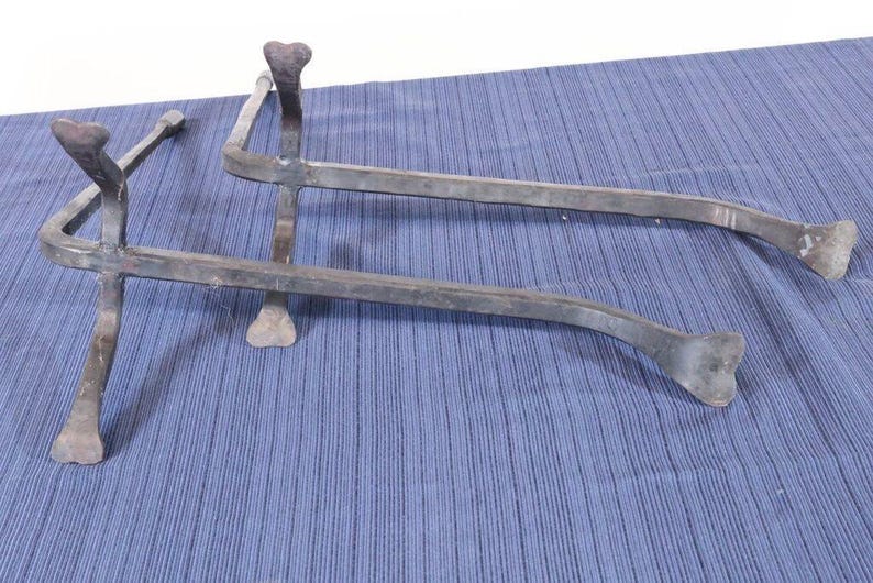 Op de afbeelding: Twee metalen trivets met een rustieke, handgemaakte uitstraling. Elke trivet heeft een gebogen, staafachtig ontwerp met hartvormige accenten aan de uiteinden en aan de bovenkant van de poten. Het metaal heeft een donkere, verouderde afwerking.