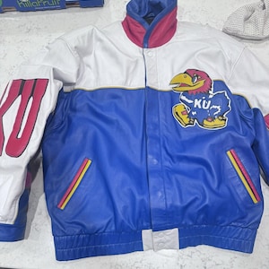 May include: A blue, white, and red leather jacket with a snap closure. The jacket features a Kansas Jayhawks logo on the chest and "KU" lettering on the sleeve. The collar is red and blue.