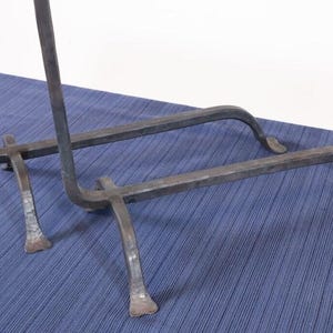 Op de afbeelding: Een donkergrijze smeedijzeren standaard. Het heeft een rechthoekige basis met gebogen steunen en een verticaal frame. De poten zijn gebogen en eindigen in een platte voet. De standaard staat op een blauw oppervlak.