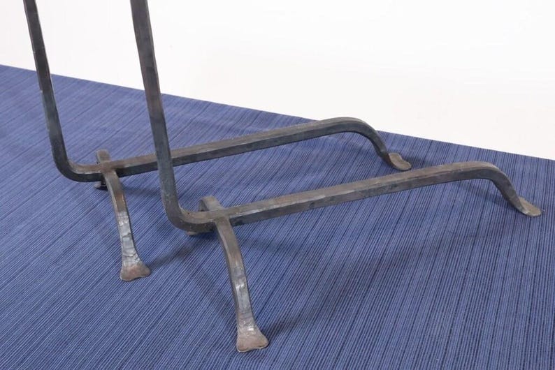 Op de afbeelding: Een donkergrijze smeedijzeren standaard. Het heeft een rechthoekige basis met gebogen steunen en een verticaal frame. De poten zijn gebogen en eindigen in een platte voet. De standaard staat op een blauw oppervlak.