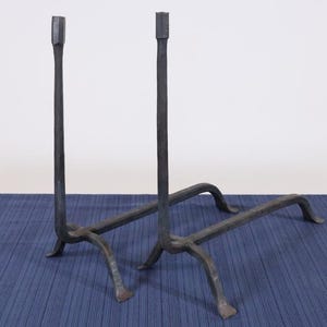 Op de afbeelding: Een paar zwarte, smeedijzeren haardijzers. Elk haardijzer heeft een lange, verticale paal met een vierkante bovenkant, ondersteund door een basis met gebogen poten. De haardijzers worden op een blauw oppervlak weergegeven.