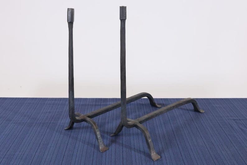Op de afbeelding: Een paar zwarte, smeedijzeren haardijzers. Elk haardijzer heeft een lange, verticale paal met een vierkante bovenkant, ondersteund door een basis met gebogen poten. De haardijzers worden op een blauw oppervlak weergegeven.