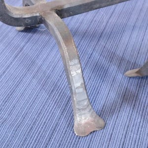 Op de afbeelding: Close-up van een smeedijzeren object met een gebogen, zwarte bovenkant en drie poten. De poten zijn lichtgrijs van kleur en hebben een getextureerd, handgesmeed uiterlijk. Het object staat op een blauw stoffen oppervlak.