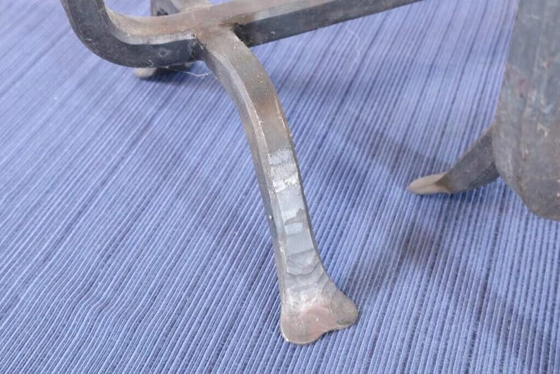 Op de afbeelding: Close-up van een smeedijzeren object met een gebogen, zwarte bovenkant en drie poten. De poten zijn lichtgrijs van kleur en hebben een getextureerd, handgesmeed uiterlijk. Het object staat op een blauw stoffen oppervlak.