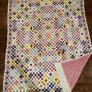 May include: A rectangular quilt featuring a multi-colored patchwork pattern of small squares in various hues including red, blue, yellow, and purple. The quilt has a white border and a folded corner revealing a pink and white floral patterned fabric backing.