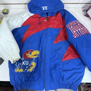 May include: A blue, red, and white hooded jacket with the text "KANSAS JAYHAWKS" on the sleeve. The jacket features a zipper closure and a graphic of the Kansas Jayhawks mascot. The jacket has a white sleeve and a blue hood.