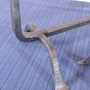 Op de afbeelding: Close-up van een handgemaakt, donker metalen object met een gebogen poot en een horizontale balk. Het metaal heeft een ruwe, getextureerde afwerking. Het object staat tegen een blauwe stoffen achtergrond.