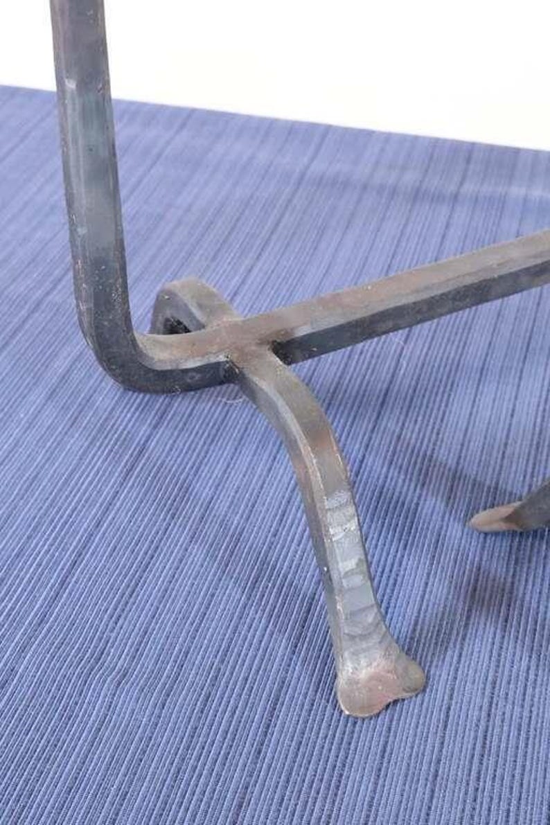 Op de afbeelding: Close-up van een handgemaakt, donker metalen object met een gebogen poot en een horizontale balk. Het metaal heeft een ruwe, getextureerde afwerking. Het object staat tegen een blauwe stoffen achtergrond.