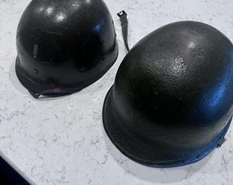 Könnte beinhalten: Zwei dunkelgrüne Militärhelme. Die Helme haben eine abgerundete Form mit einem Rand um die Basis. Ein Helm hat einen Riemen befestigt. Die Helme scheinen aus Metall zu sein und haben eine matte Oberfläche.