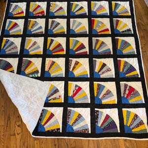 May include: A quilt featuring a grid of square blocks, each containing a multi-colored fan pattern with various floral and geometric prints. The blocks are separated by black sashing, and the quilt has a white backing visible at one corner.