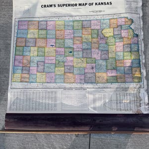 May include: A vintage map of Kansas, titled "Cram's Superior Map of Kansas," is displayed. The map is rolled up at the bottom and has a wooden rod. The map features a colorful grid of counties and detailed geographical information.