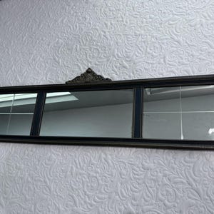 May include: A rectangular mirror with a dark frame and three mirrored sections. The frame has a decorative top piece and a beaded edge. The mirror is long and horizontal, suitable for a wall.
