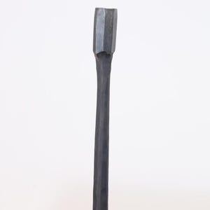 Op de afbeelding: Een donkergrijze, met de hand gesmede metalen tool met een rechthoekige kop en een lange, slanke steel. De tool wordt waarschijnlijk gebruikt voor het vormen of bewerken van metaal of hout. De achtergrond is een effen witte muur.