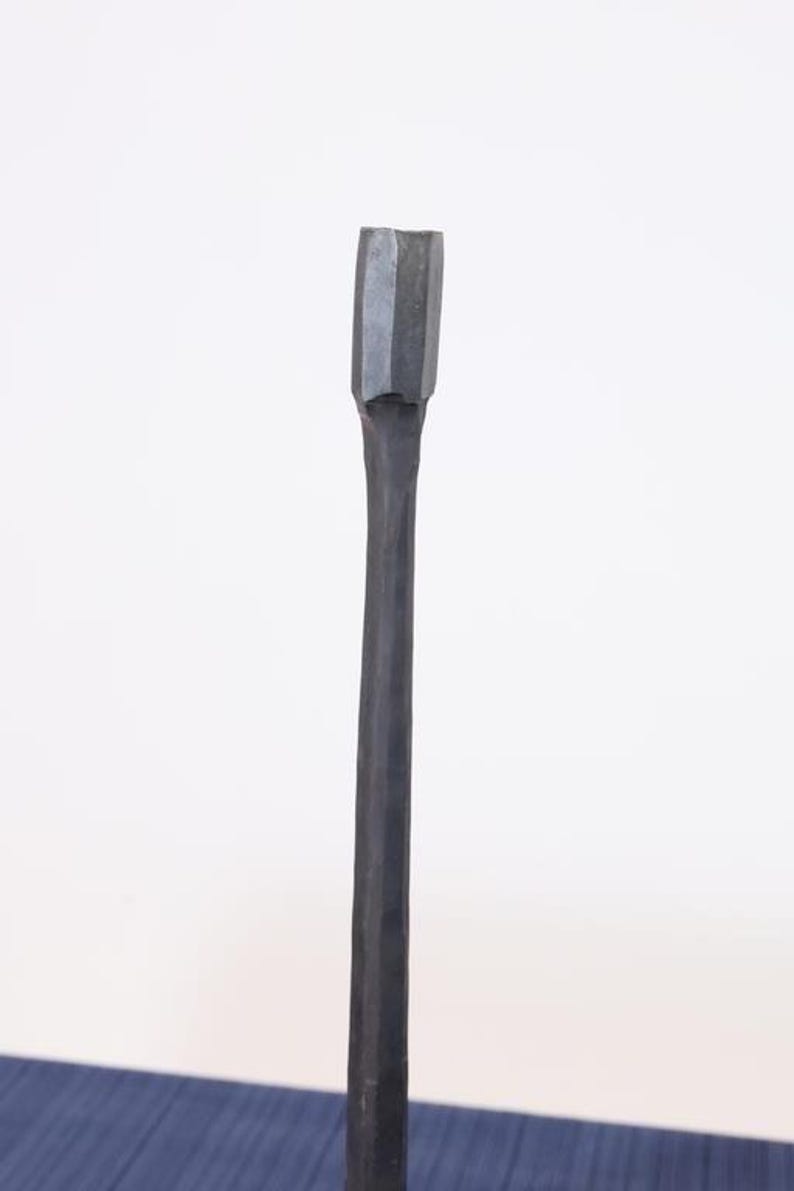 Op de afbeelding: Een donkergrijze, met de hand gesmede metalen tool met een rechthoekige kop en een lange, slanke steel. De tool wordt waarschijnlijk gebruikt voor het vormen of bewerken van metaal of hout. De achtergrond is een effen witte muur.