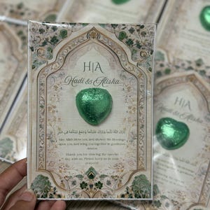 May include: A wedding favor card with a green heart-shaped chocolate. The card has an ornate design with the names "Hadi & Alisha" and text in English and Arabic. The card is rectangular with a decorative border.