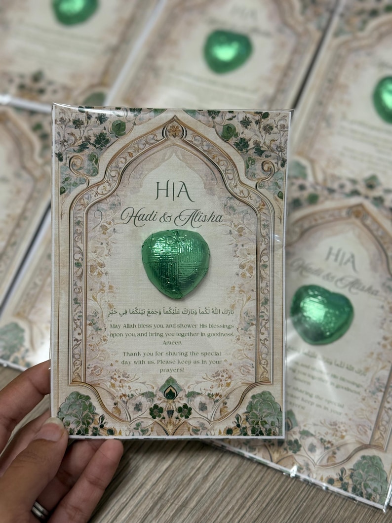 May include: A wedding favor card with a green heart-shaped chocolate. The card has an ornate design with the names "Hadi & Alisha" and text in English and Arabic. The card is rectangular with a decorative border.