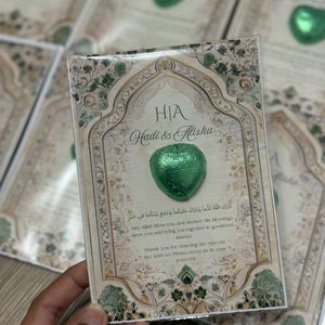 May include: A wedding favor card with a green heart-shaped chocolate. The card has an ornate design with the names "Hadi & Alisha" and text in English and Arabic. The card is held in a hand.