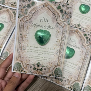 May include: A stack of wedding favor cards with a green foil-wrapped heart-shaped chocolate. The cards feature ornate floral designs, the names "Hadi & Alisha", and text in English and Arabic. The cards are held by a hand.