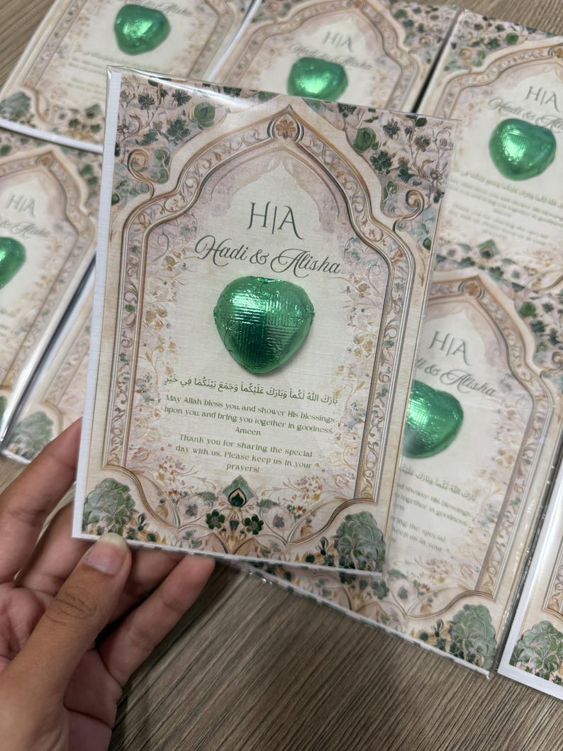 May include: A stack of wedding favor cards with a green foil-wrapped heart-shaped chocolate. The cards feature ornate floral designs, the names "Hadi & Alisha", and text in English and Arabic. The cards are held by a hand.