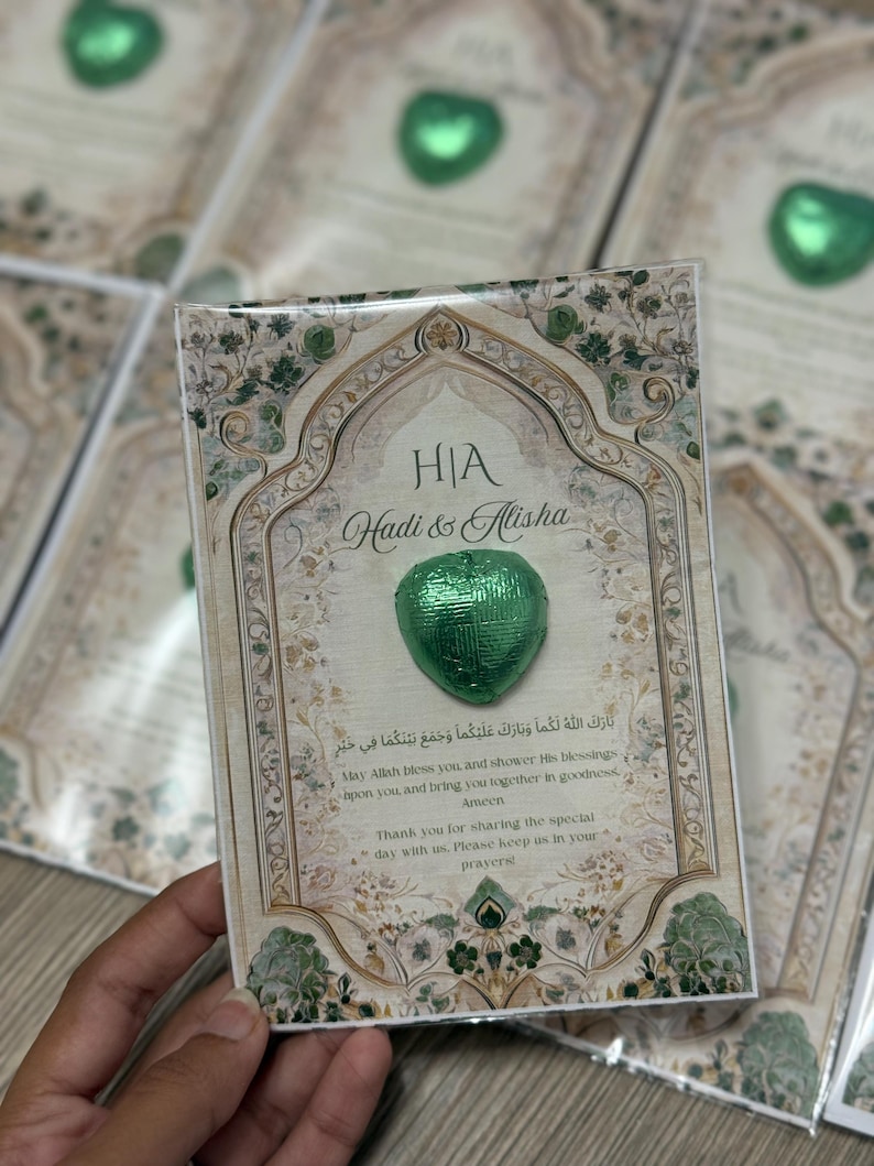 May include: Wedding favor card with a green heart-shaped chocolate. The card has a beige background with green floral accents and text in English and Arabic. The names "Hadi & Alisha" are printed on the card.