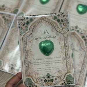 May include: A wedding favor card with a green foil-wrapped heart-shaped chocolate. The card features an ornate design with green and beige tones, the names "Hadi & Alisha", and a religious blessing in Arabic and English.