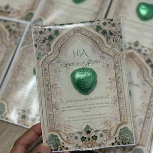 May include: A wedding favor card with a green heart-shaped chocolate. The card has an ornate design with the names "Hadi & Alisha" and text in English and Arabic. The card is held in a hand.